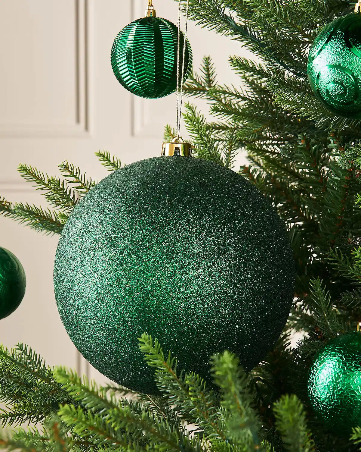 Oversized Christmas Green glittery Christmas ball ornament with a gold hook on a Christmas tree branch against a cream panel background with 4 smaller Green baubles on branch