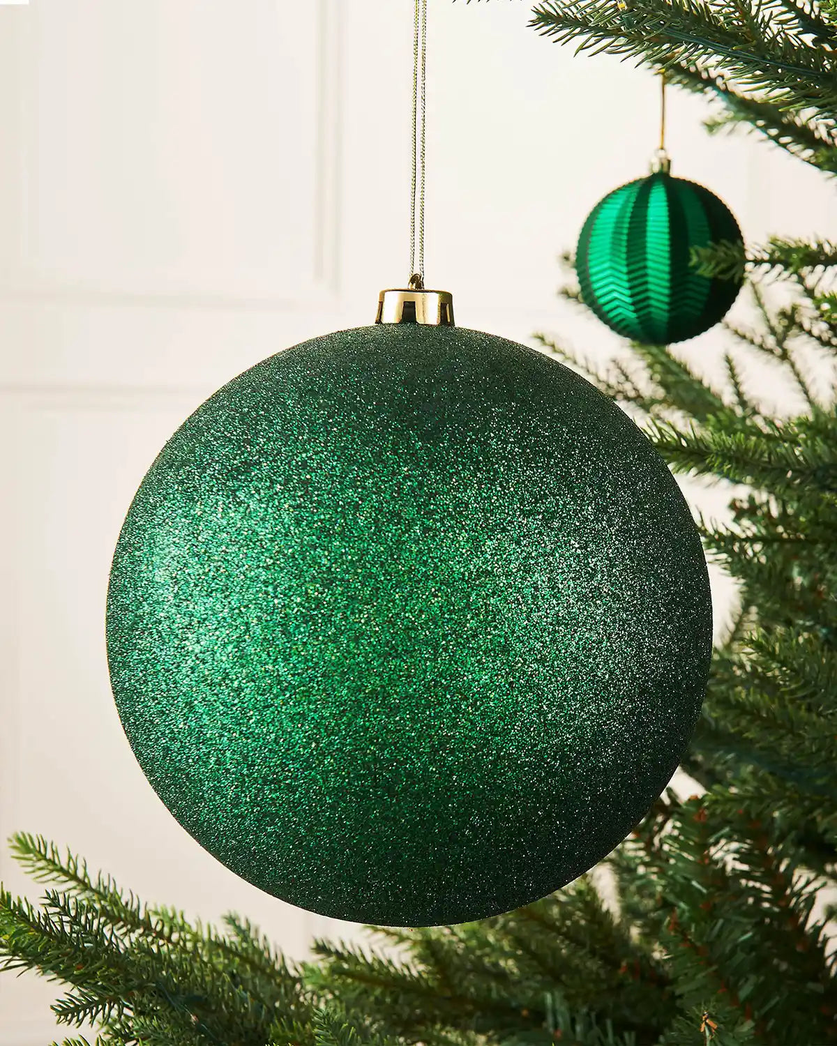 Oversized Christmas Green glittery Christmas ball ornament with a gold hook on a Christmas tree branch against a cream panel background with a smaller Green bauble on branch