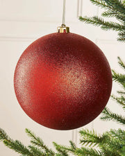 Oversized Red glittery Christmas bauble ornament hanging on a tree branch against a cream panel wall
