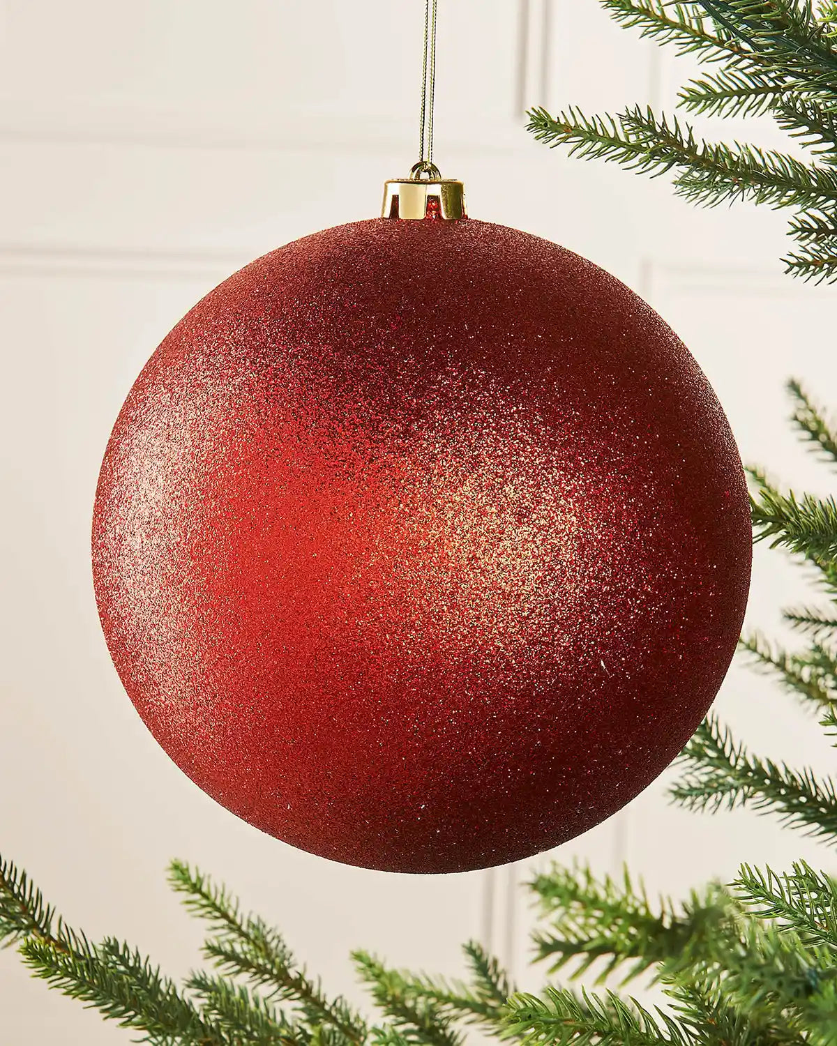 Oversized Red glittery Christmas bauble ornament hanging on a tree branch against a cream panel wall