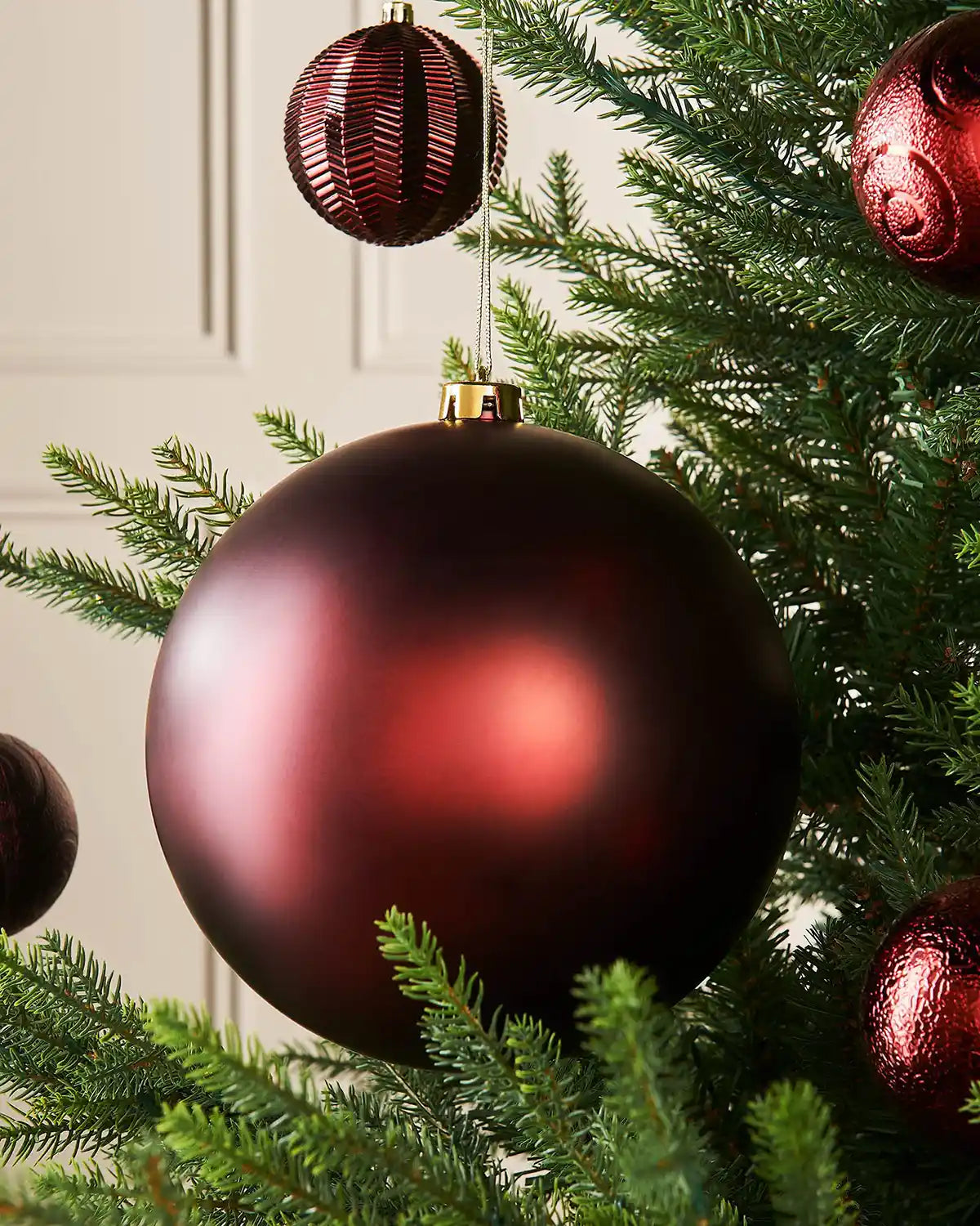 Oversized burgundy matt bauble on a Christmas tree branch with 4 smaller burgundy baubles in the background