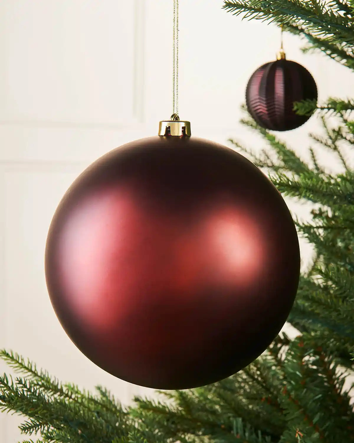 Oversized burgundy matt bauble on a Christmas tree branch with a smaller burgundy bauble in the background