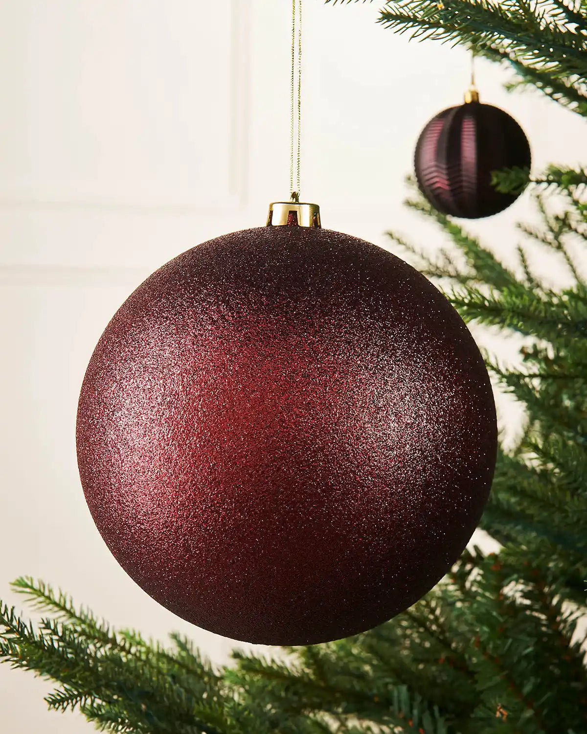 Oversized glitter Burgundy bauble on a Christmas tree branch with a smaller burgundy bauble in background