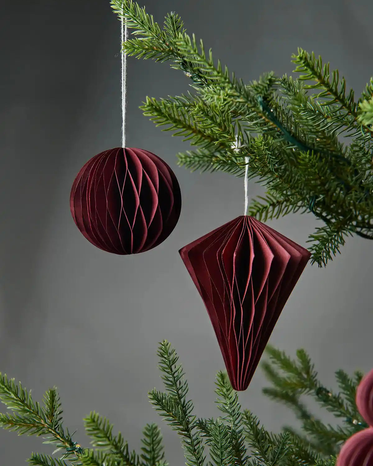 A pair of 10cm burgundy red paper Christmas tree decorations hanging on a tree in front of a grey wall background