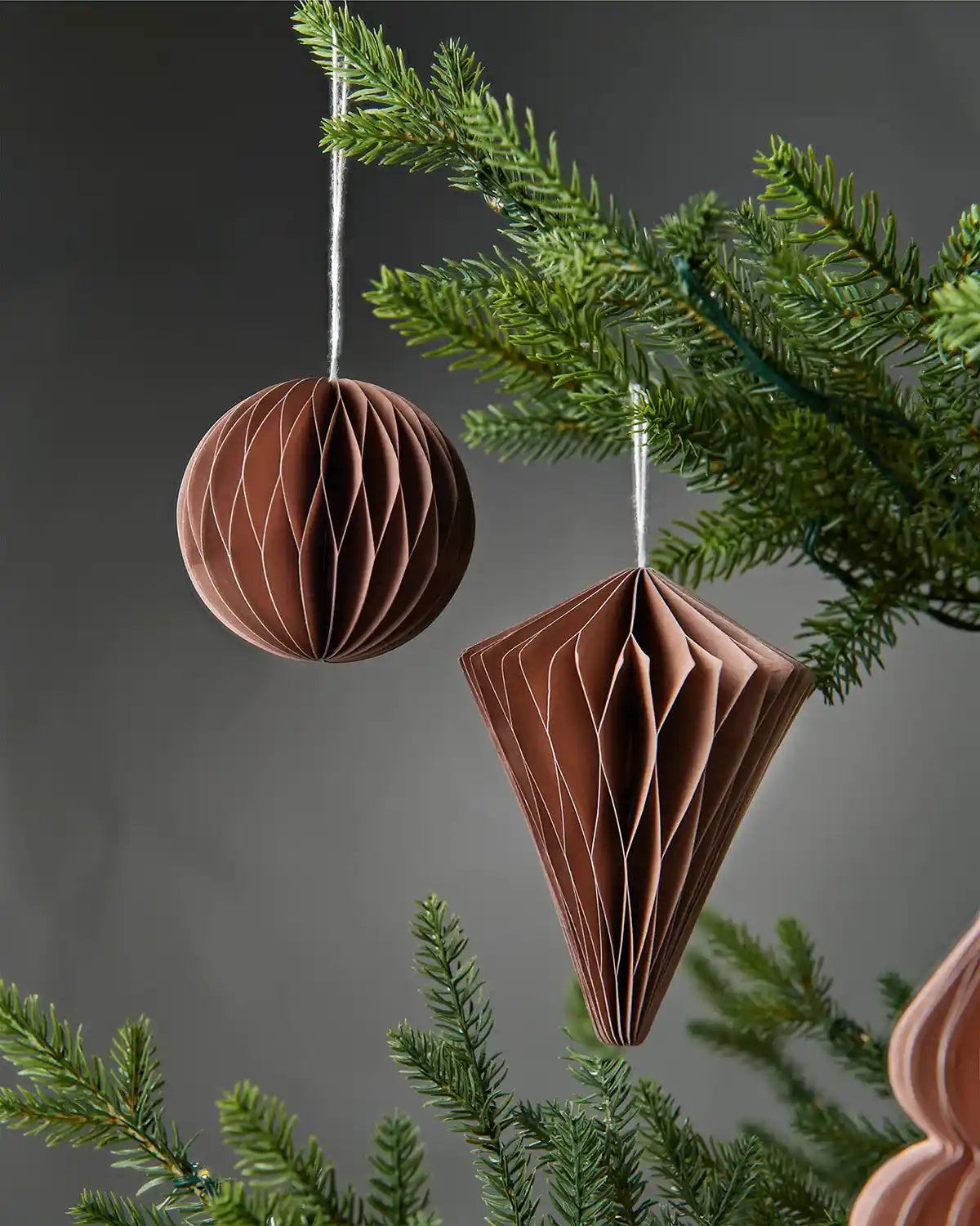A pair of 10cm mocha brown paper Christmas decorations hanging on a tree in front of a grey wall background