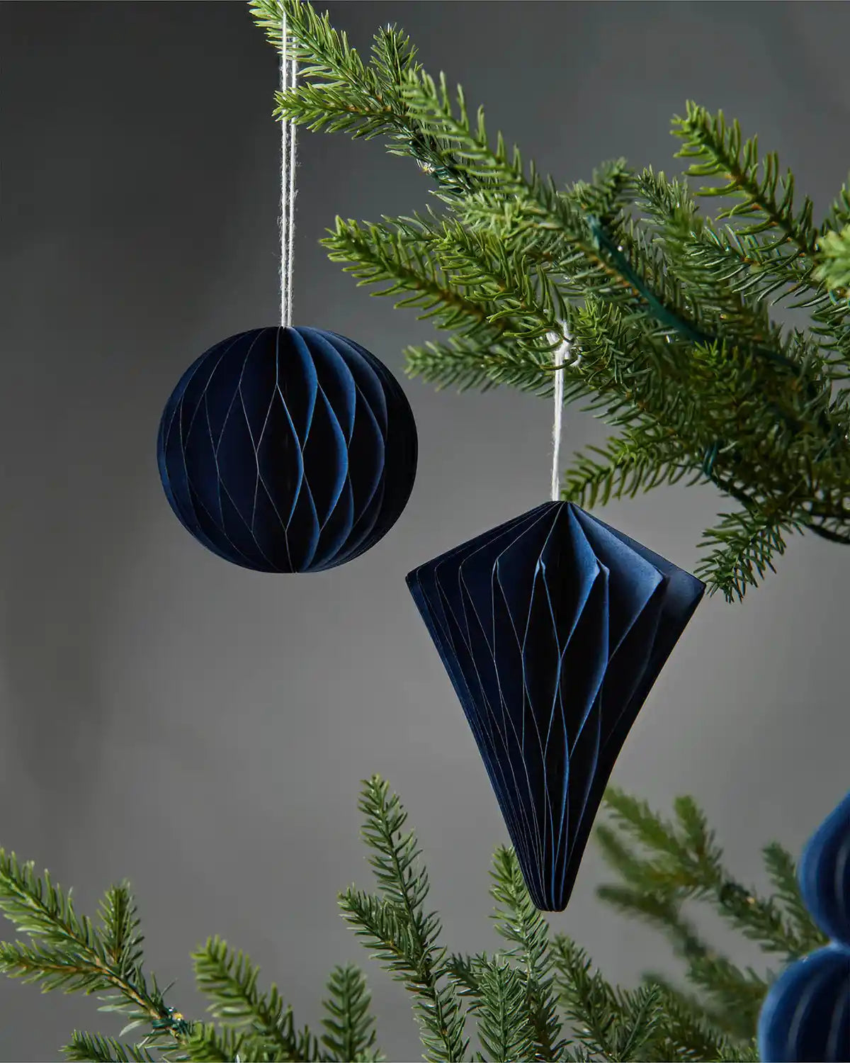 Pair of 10cm navy blue paper honeycomb tree decorations hanging on Christmas tree in front of grey wall background