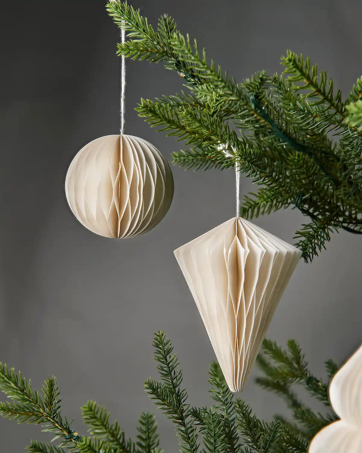 A pair of 10cm off-white paper Christmas tree decorations hanging on branches in front of grey wall background