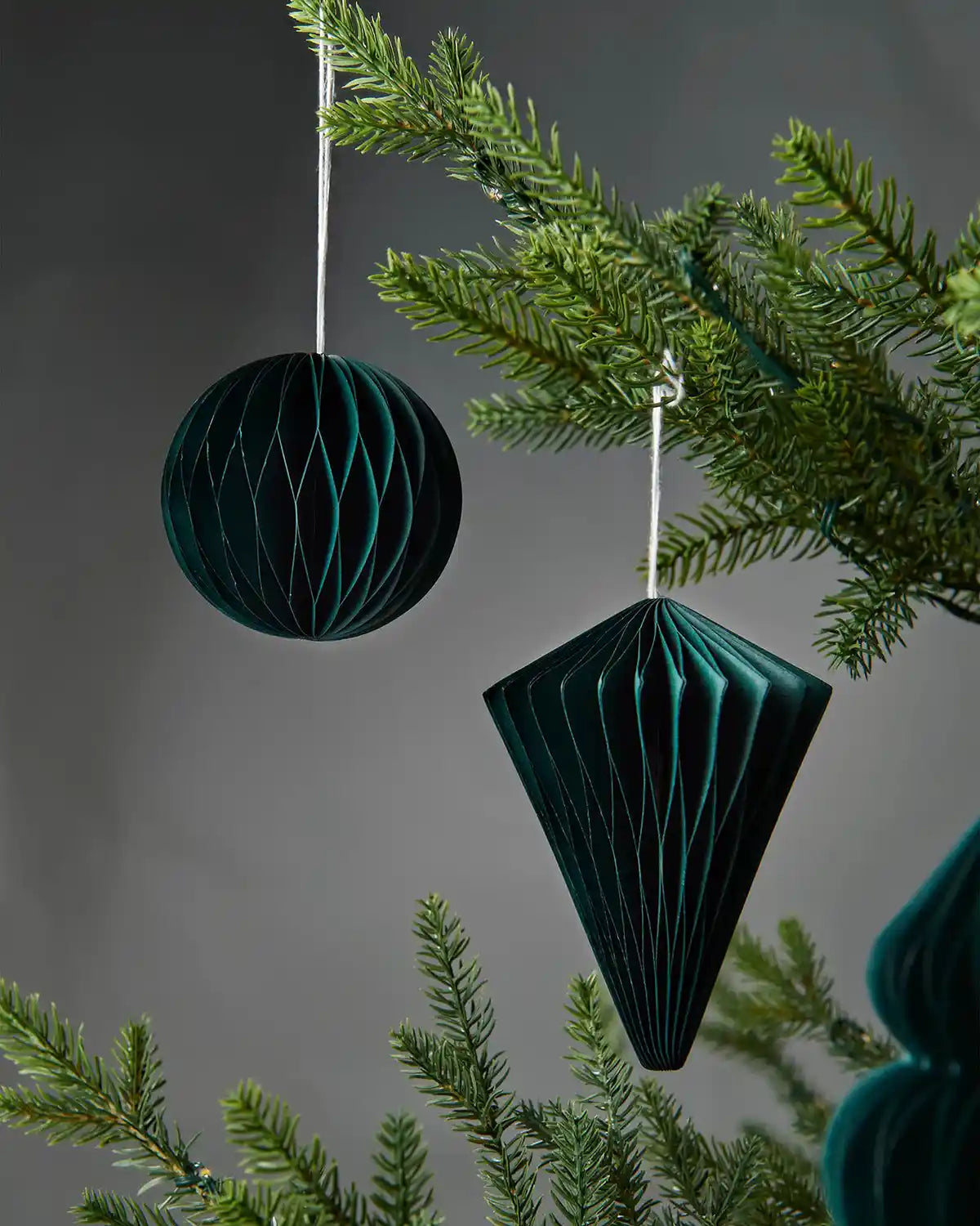 A pair of 10cm sage green paper honeycomb tree decorations hanging on Christmas tree in front of grey wall background