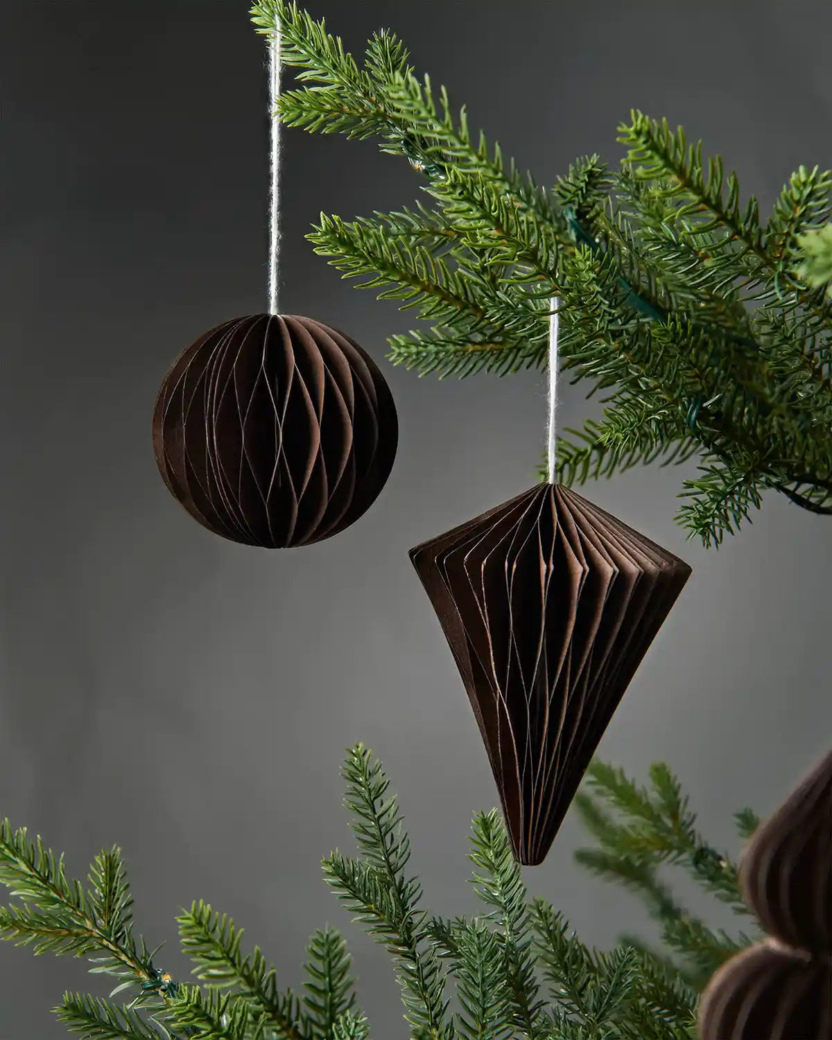 Pair of espresso brown paper honeycomb tree decorations hanging on Christmas tree in front of grey wall background