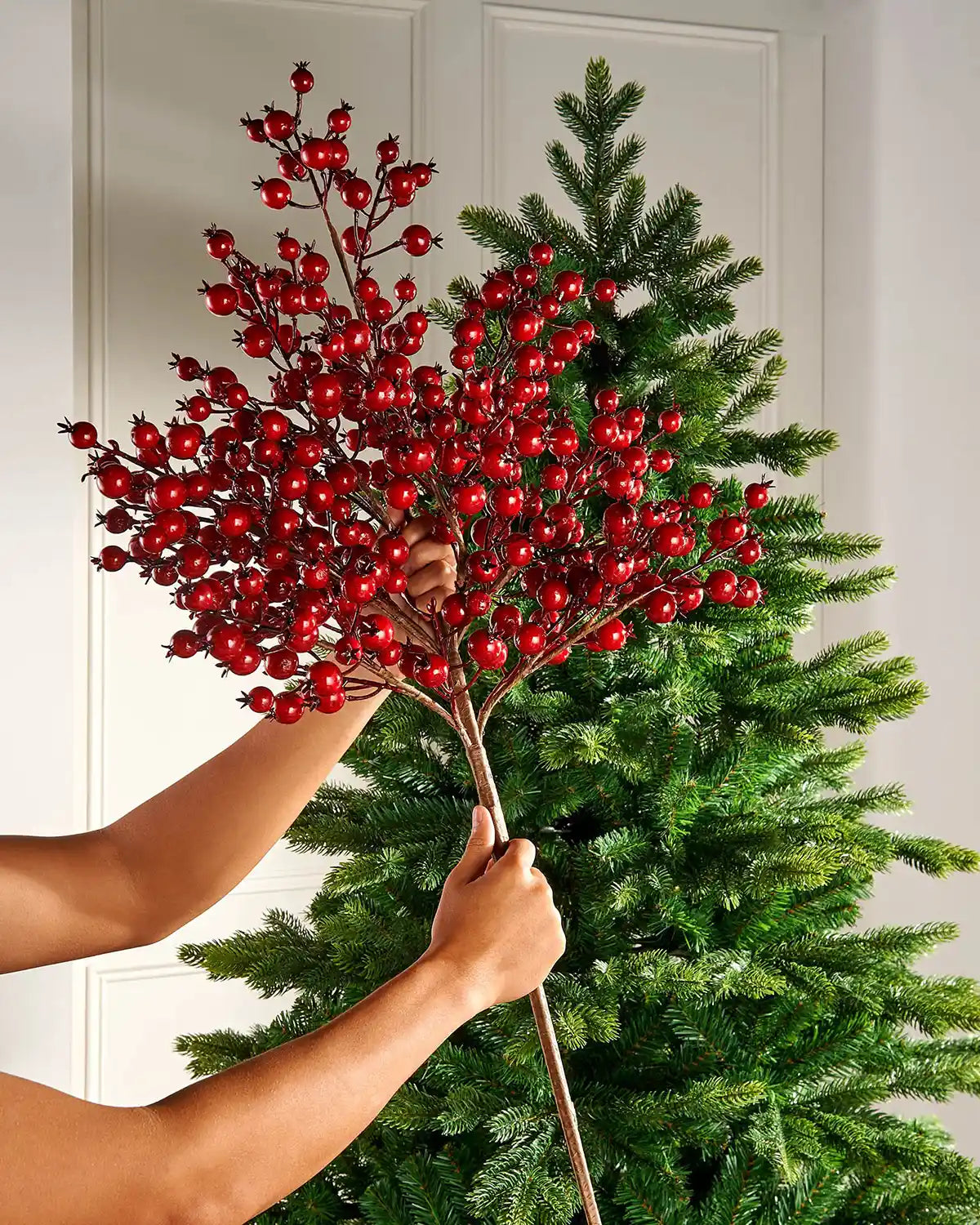 Person holding a red berry branch in front of a Christmas tree.