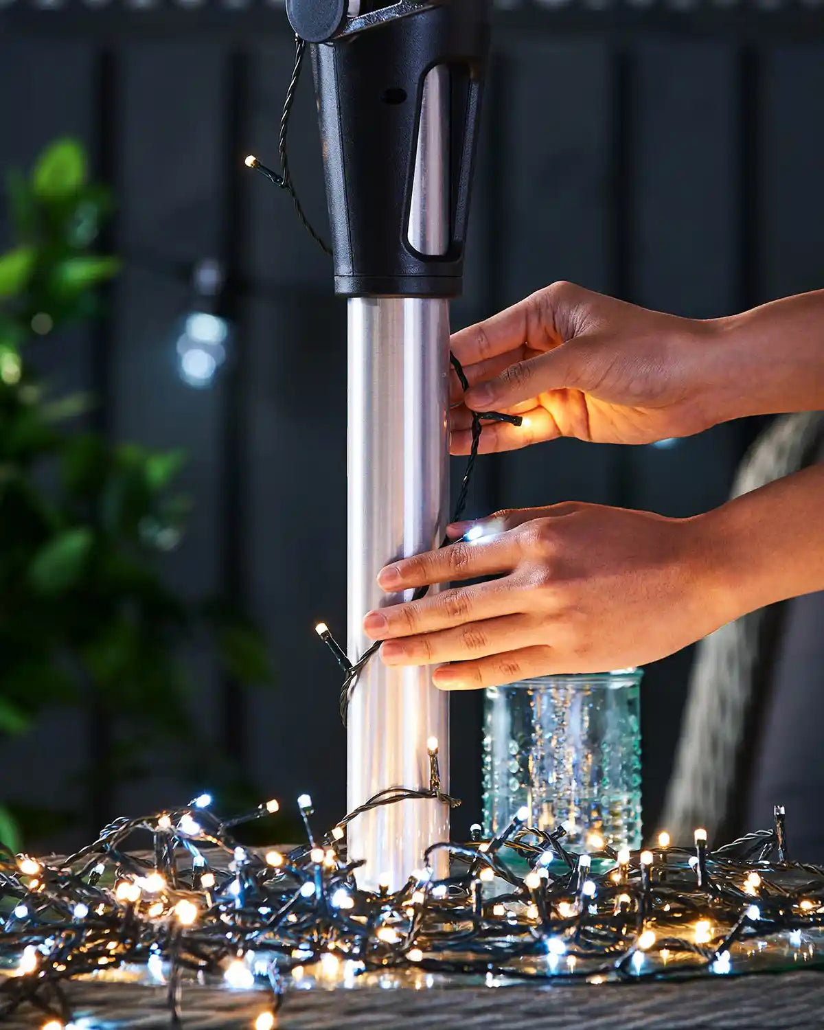 Person stringing warm white and ice white lights on an outdoor umbrella pole with a blurred background