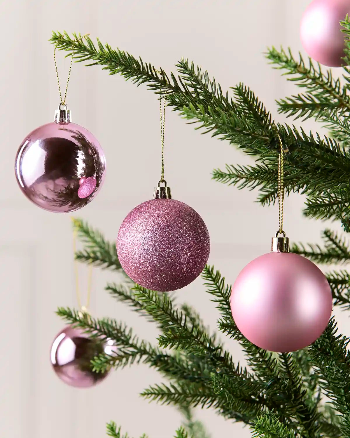 Pink Christmas tree baubles hanging on green pine branches, including a glittery sphere and shiny reflective balls against a soft light background.