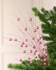 Pink glittery berry branches with sparkling round ornaments, styled among green pine needles against a cream paneled wall.
