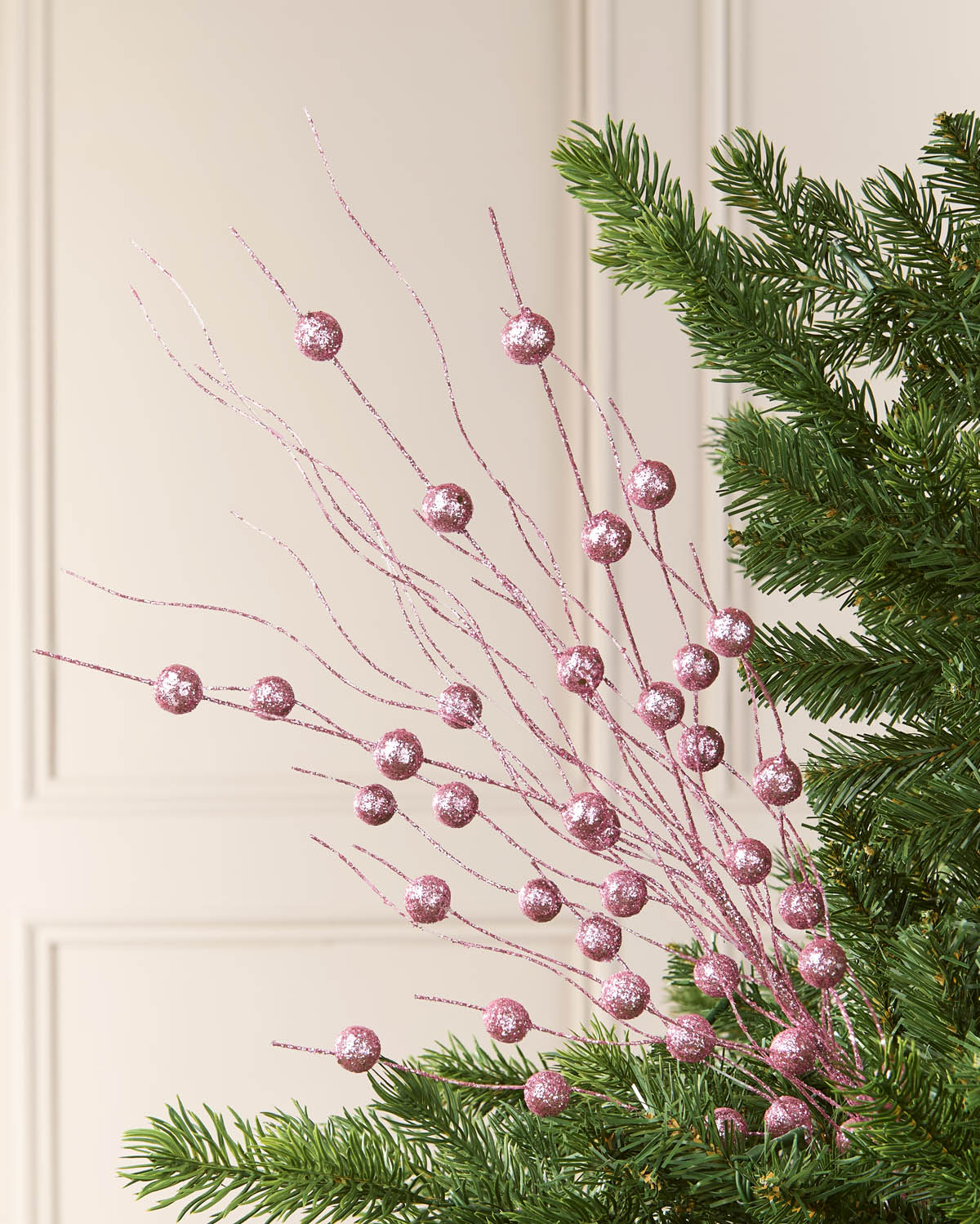 Pink glittery berry branches with sparkling round ornaments, styled among green pine needles against a cream paneled wall.