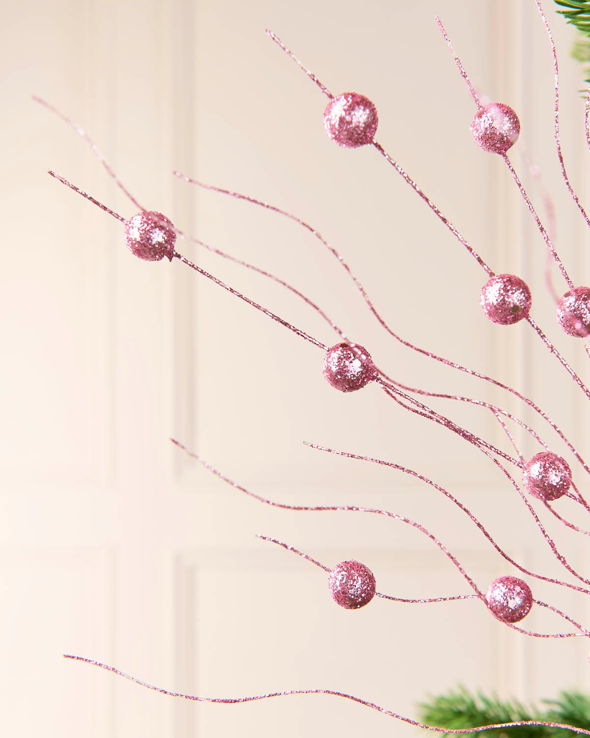 Pink glittery decorative branches with round pom-pom ornaments, shown against a soft beige paneled background.