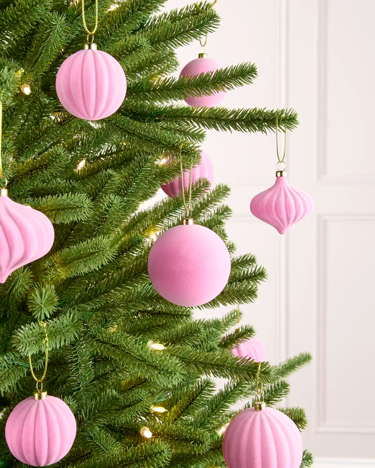 Pink ribbed and smooth Christmas tree ornaments hanging on a green pine tree with warm white string lights, set against a white panneled wall.