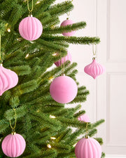 Pink ribbed and smooth Christmas tree ornaments hanging on a green pine tree with warm white string lights, set against a white panneled wall.