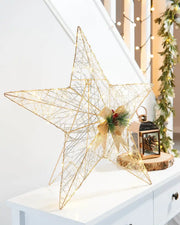 Side view of Pre-Lit 2D Metal Star, Gold, 60 cm on a White table with Pinecone lantern in front of a staircase with lights and a garland  
