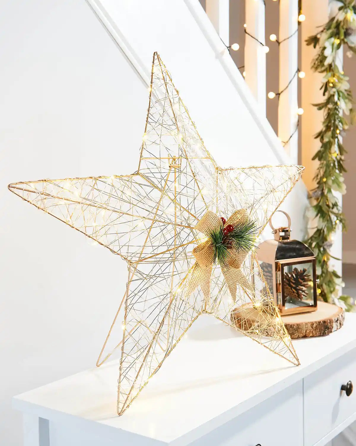 Side view of Pre-Lit 2D Metal Star, Gold, 60 cm on a White table with Pinecone lantern in front of a staircase with lights and a garland  