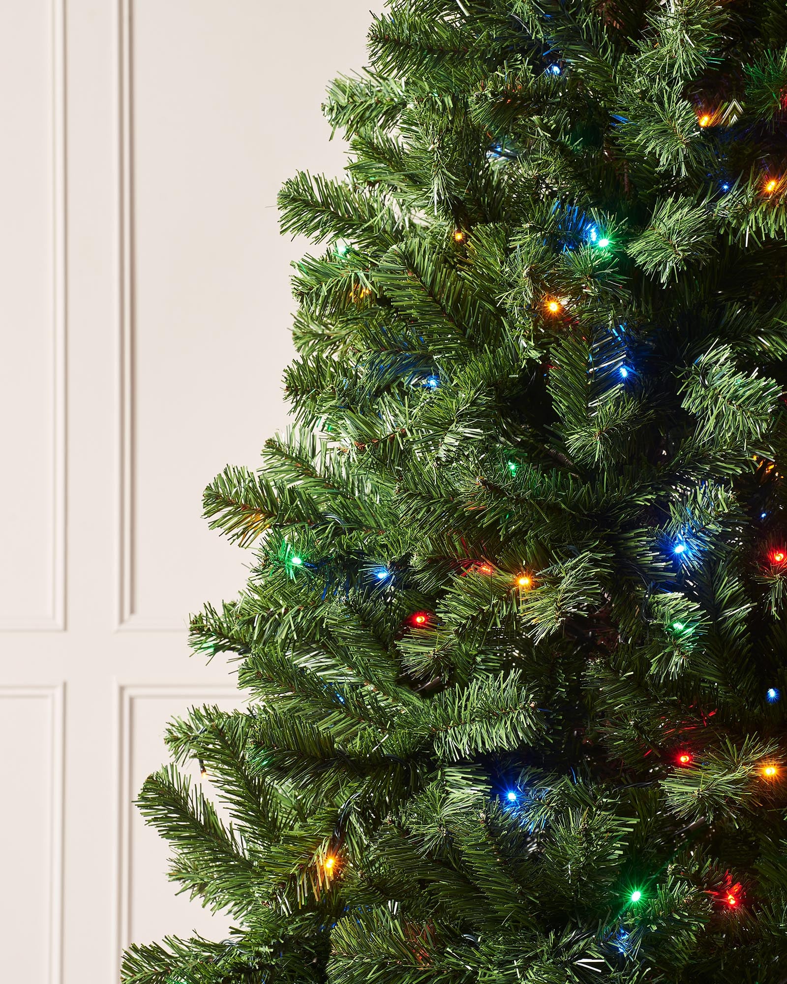 Pre-lit Emerald Spruce artificial Christmas tree with realistic PVC branches and multi-coloured LED lights featuring 8 lighting functions, displayed indoors against a white wall and wooden floor.