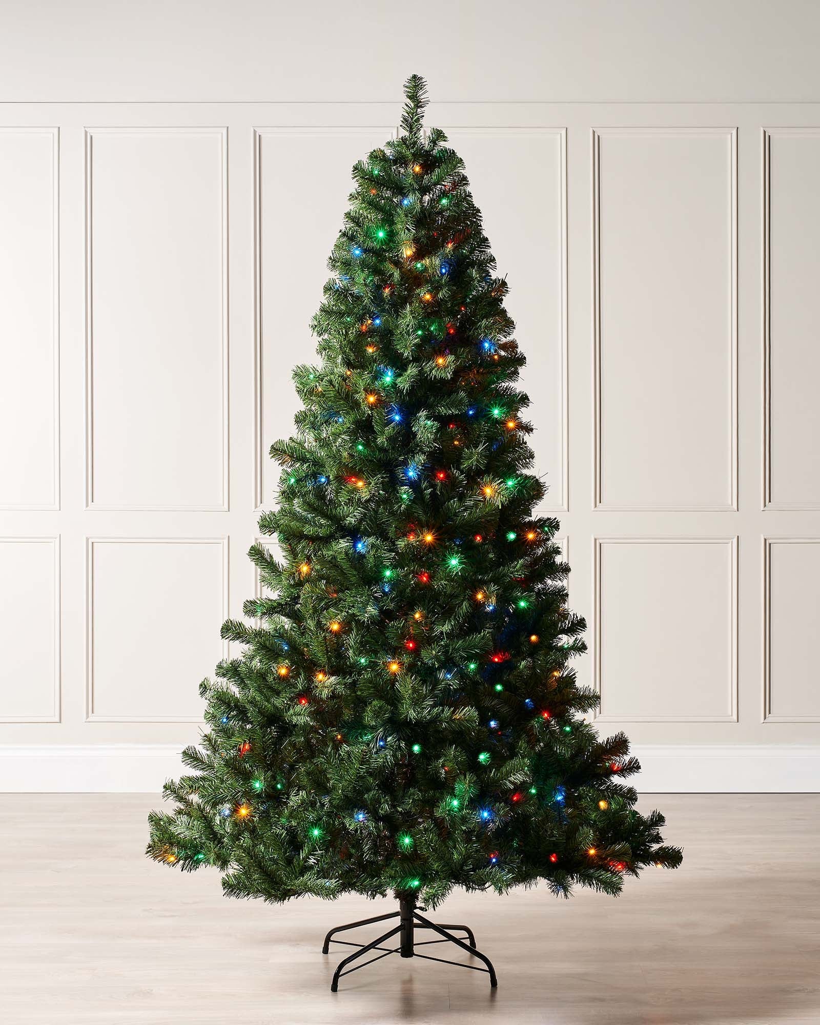 Pre-lit Emerald Spruce artificial Christmas tree with realistic PVC branches and multi-coloured LED lights featuring 8 lighting functions, displayed indoors against a white wall and wooden floor.