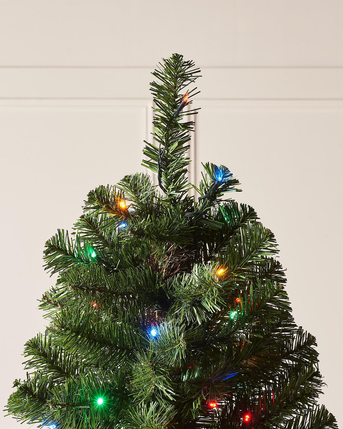 Pre-lit Emerald Spruce artificial Christmas tree with realistic PVC branches and multi-coloured LED lights featuring 8 lighting functions, displayed indoors against a white wall and wooden floor.
