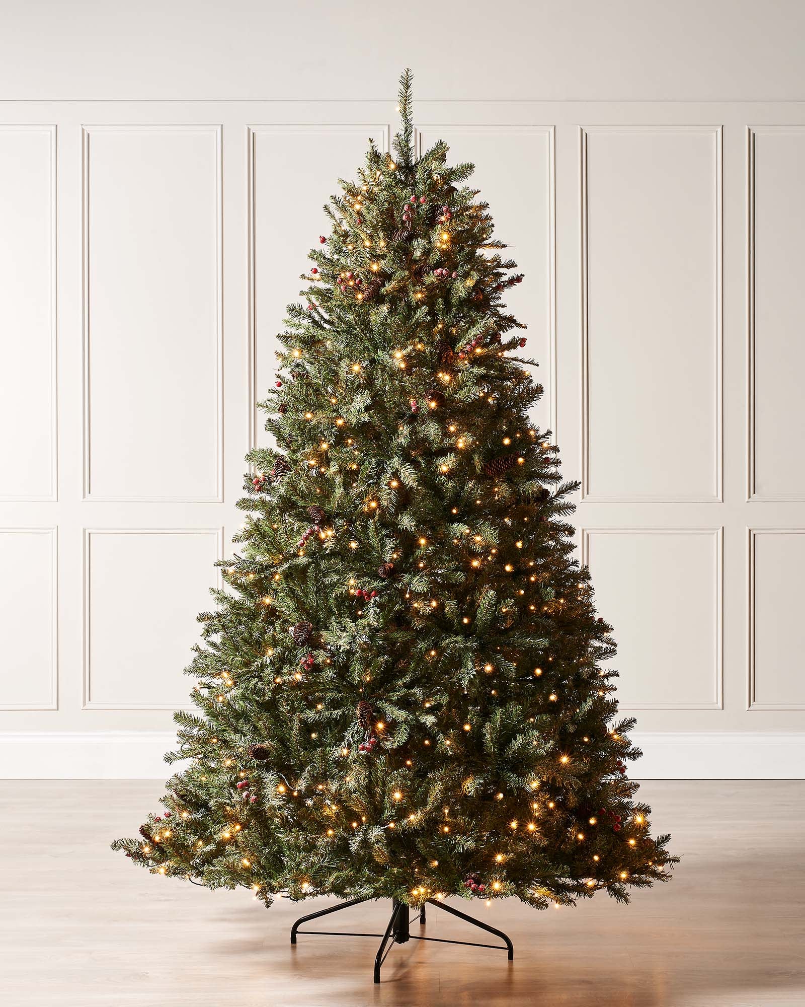 Pre-lit full-bodied artificial Christmas tree with snow-flocked branches, red berries, pinecones, and warm white LED lights, set indoors against a white wall.