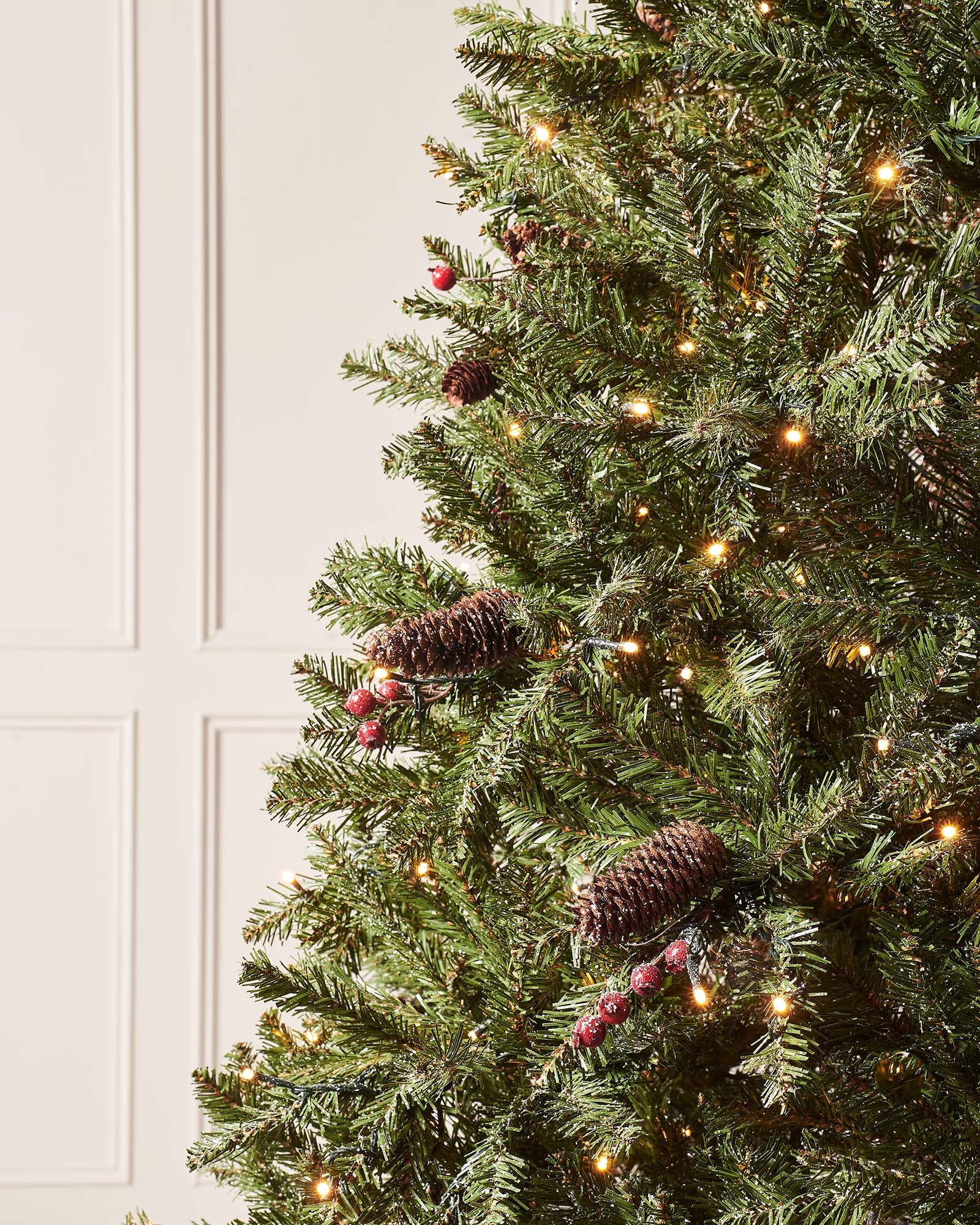 Pre-lit full-bodied artificial Christmas tree with snow-flocked branches, red berries, pinecones, and warm white LED lights, set indoors against a white wall.