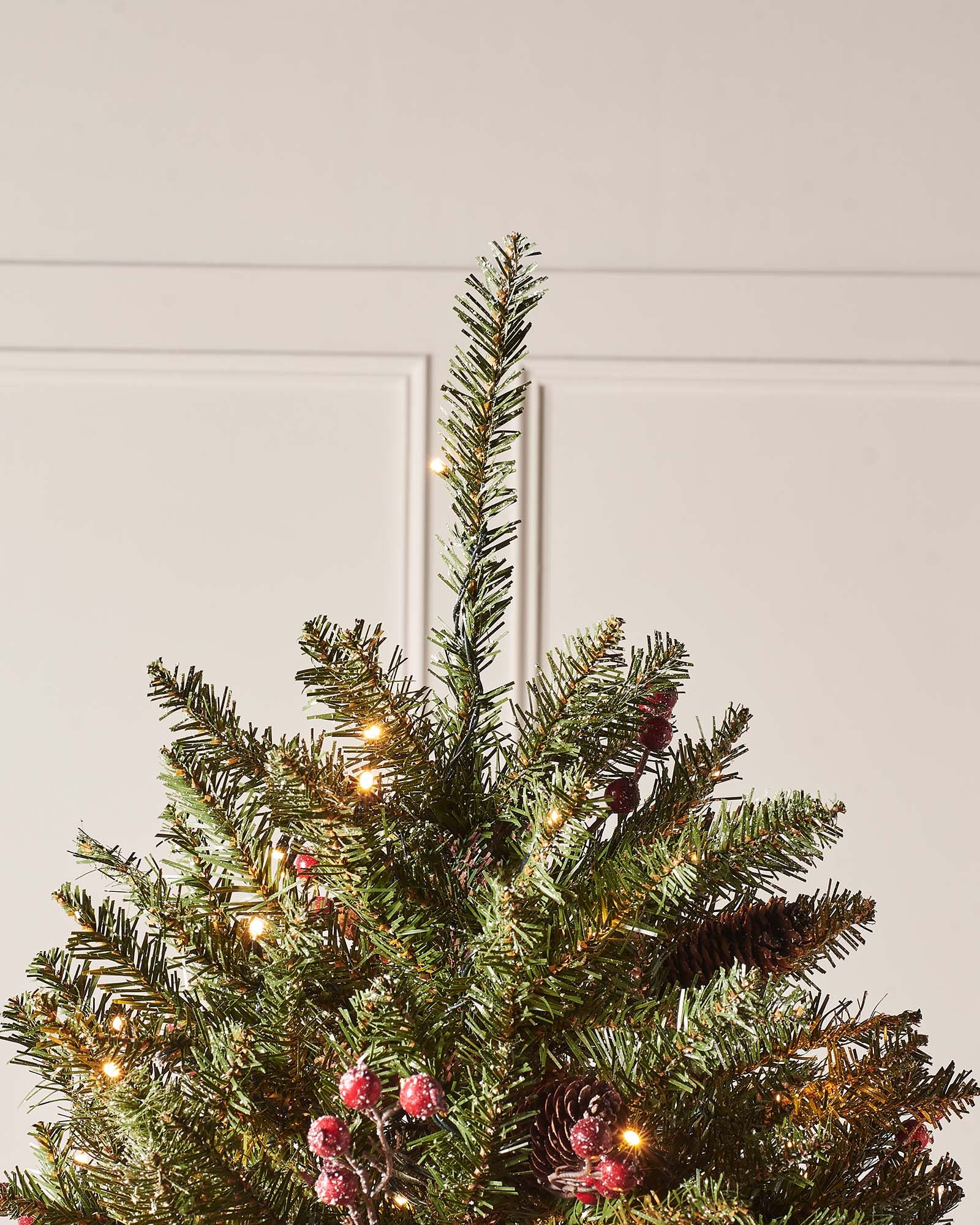 Pre-lit full-bodied artificial Christmas tree with snow-flocked branches, red berries, pinecones, and warm white LED lights, set indoors against a white wall.