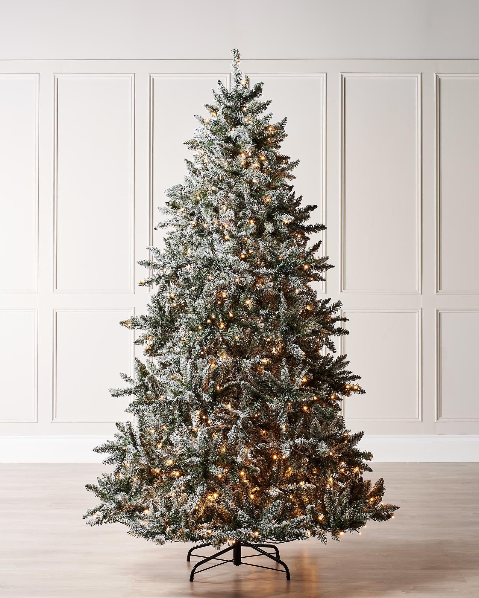 Pre-lit, full-bodied snow-flocked artificial Christmas tree with warm white LED lights, set in a room with white wall paneling.