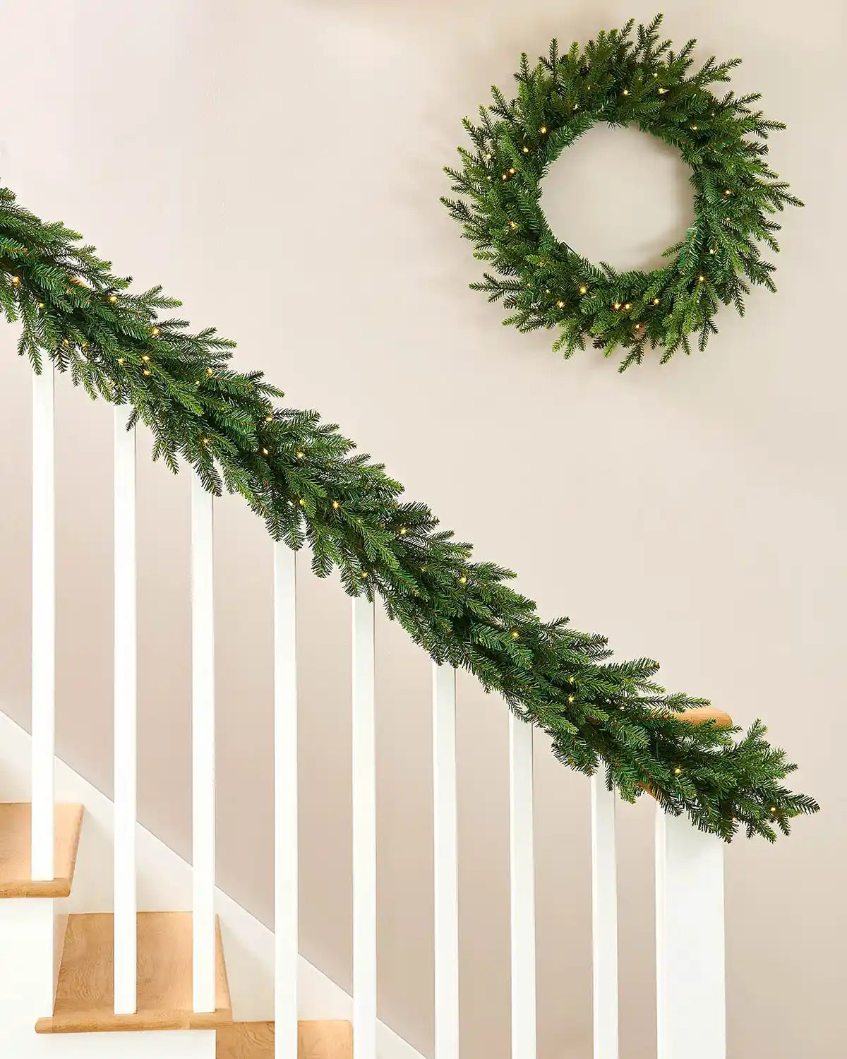 Pre-Lit Multi-Function Douglas Fir Garland, 9 ft on a wooden staircase banister and a cream wall with a matching Pre-Lit Multi Function Douglas Fir Wreath on wall.