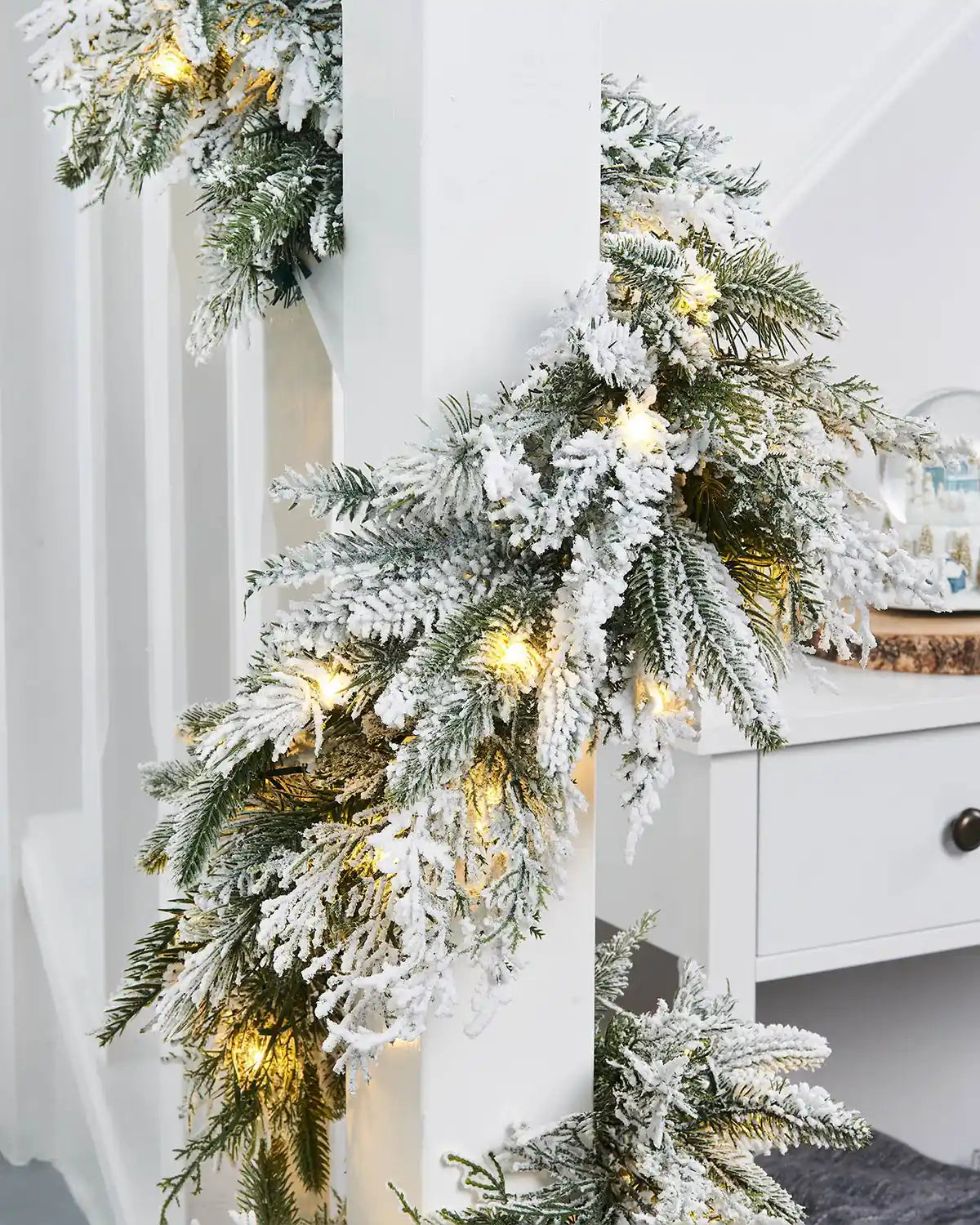 Pre-Lit Snow-Flocked Fir Garland, 18 ft on White wooden staircase banister with a snow globe on a table 