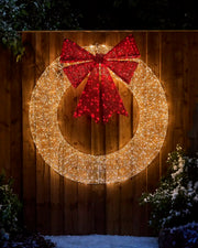 Pre-Lit Twinkling Wreath Silhouette glowing with warm white lights and a large red glitter bow, displayed outdoors against a wooden fence