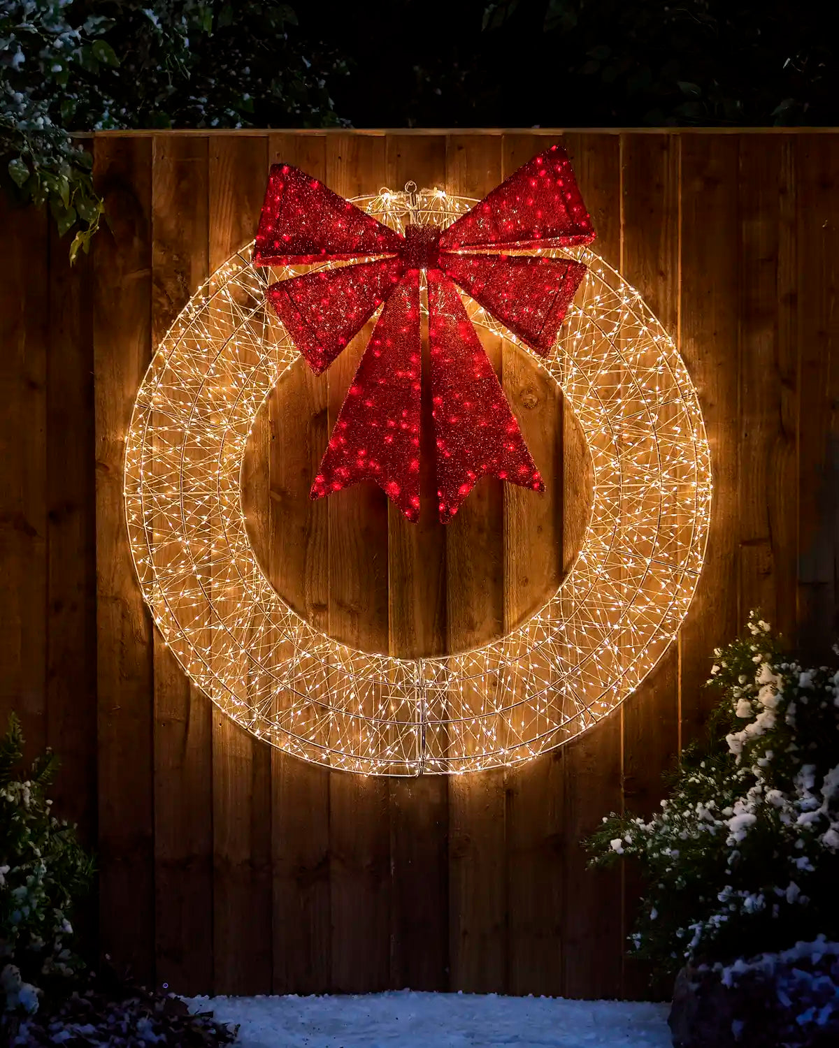 Pre-Lit Twinkling Wreath Silhouette glowing with warm white lights and a large red glitter bow, displayed outdoors against a wooden fence