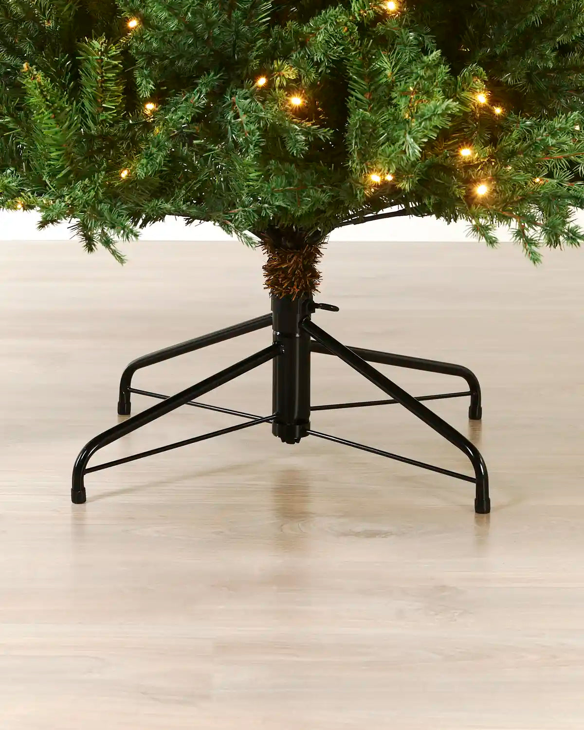 A black metal Christmas tree stand supporting a green artificial Christmas tree with warm white string lights, shown on a light wood floor.