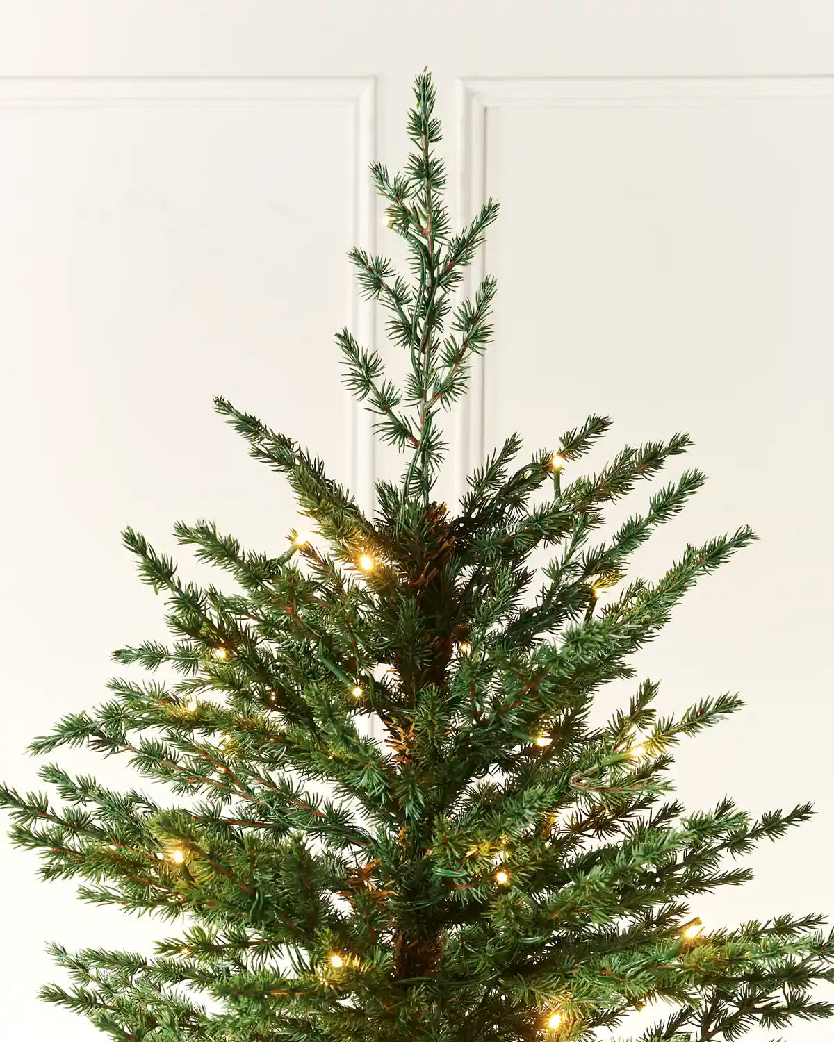 A close-up of a green Christmas tree with warm white string lights glowing through the branches, set against a clean white paneled wall.