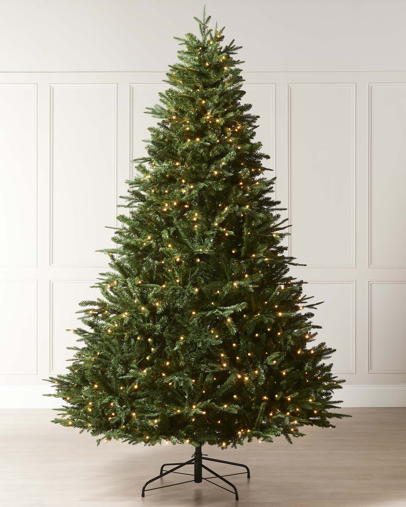 Pre-lit Grand Alaskan Fir artificial Christmas tree with warm white LED lights, full mixed PE and PVC branches, displayed against a white paneled wall.
