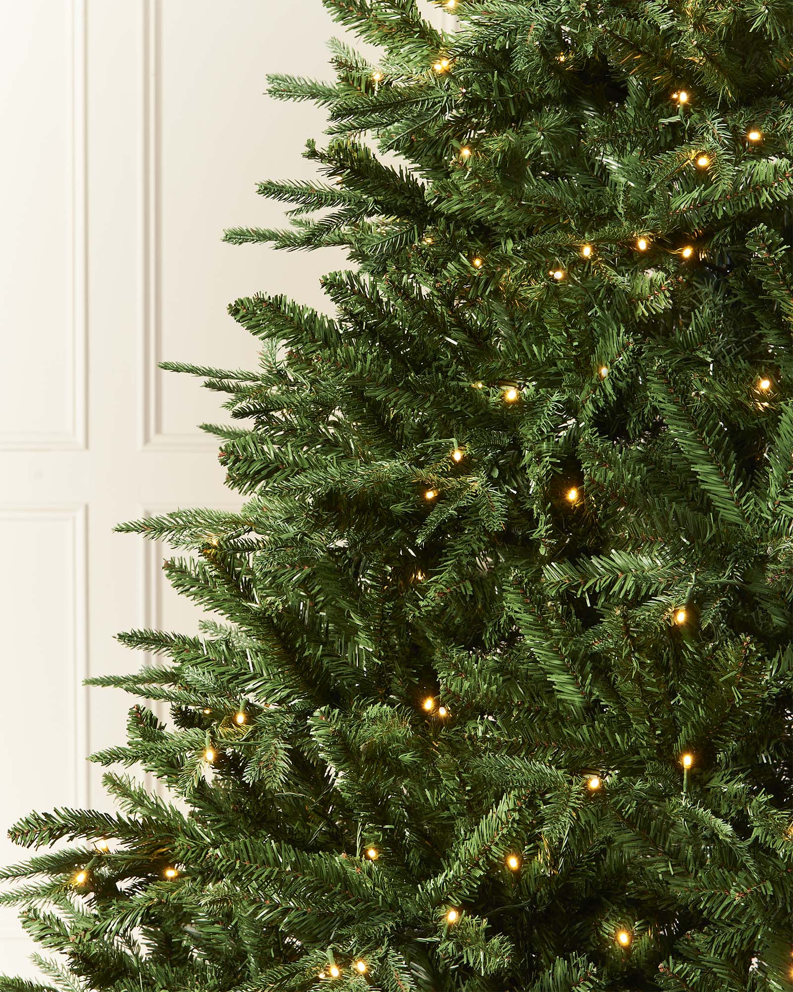 Pre-lit Grand Alaskan Fir artificial Christmas tree with warm white LED lights, full mixed PE and PVC branches, displayed against a white paneled wall.