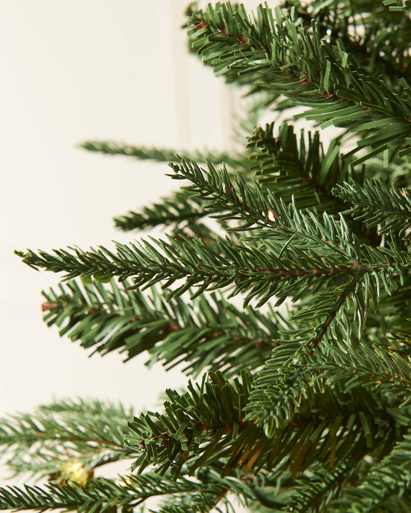 Close-up of green artificial Christmas tree branches on a light background. Pre-lit Grand Alaskan Fir artificial Christmas tree with warm white LED lights, full mixed PE and PVC branches, displayed against a white paneled wall.