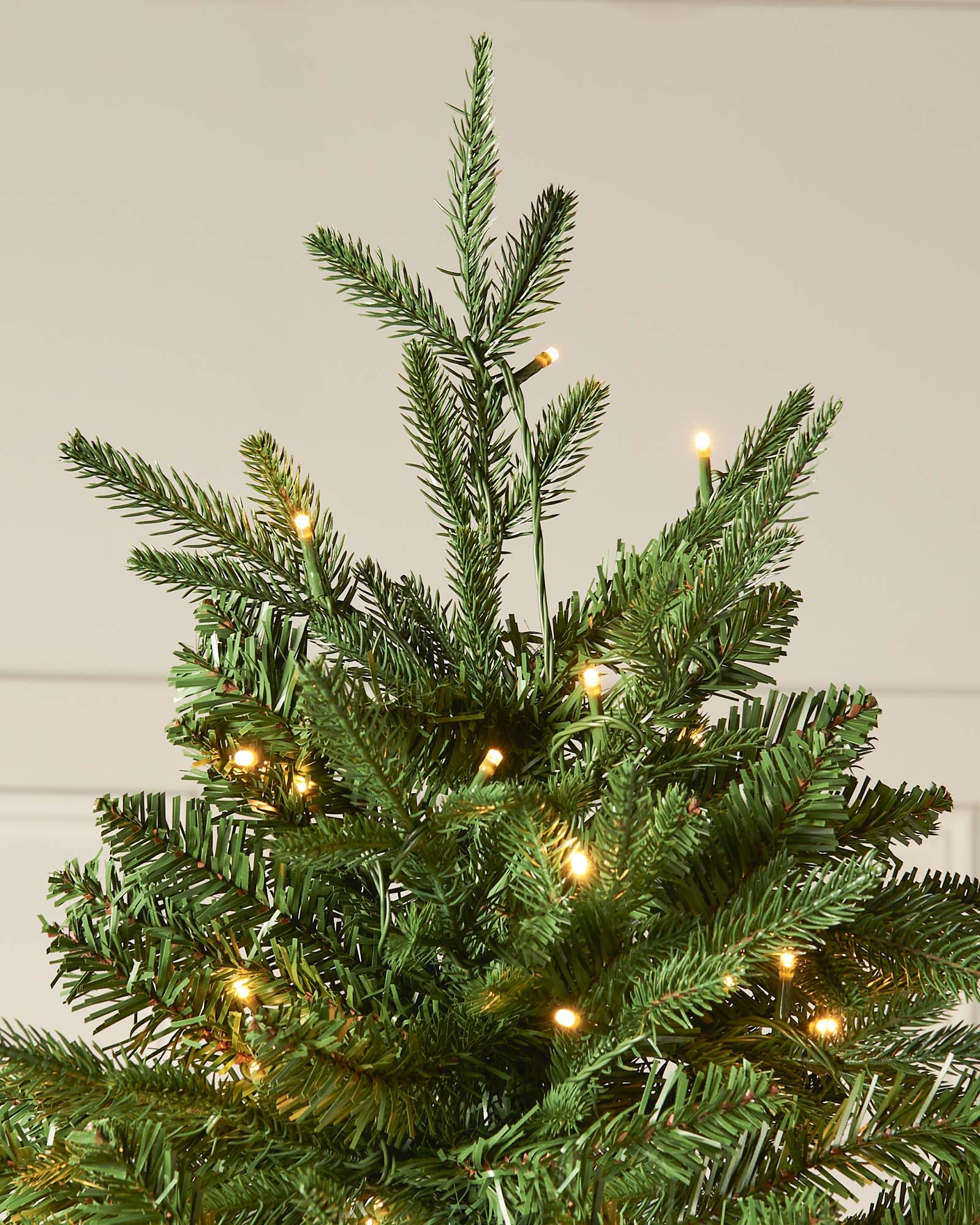 Small artificial Christmas tree with lights against a neutral background. Pre-lit Grand Alaskan Fir artificial Christmas tree with warm white LED lights, full mixed PE and PVC branches, displayed against a white paneled wall.