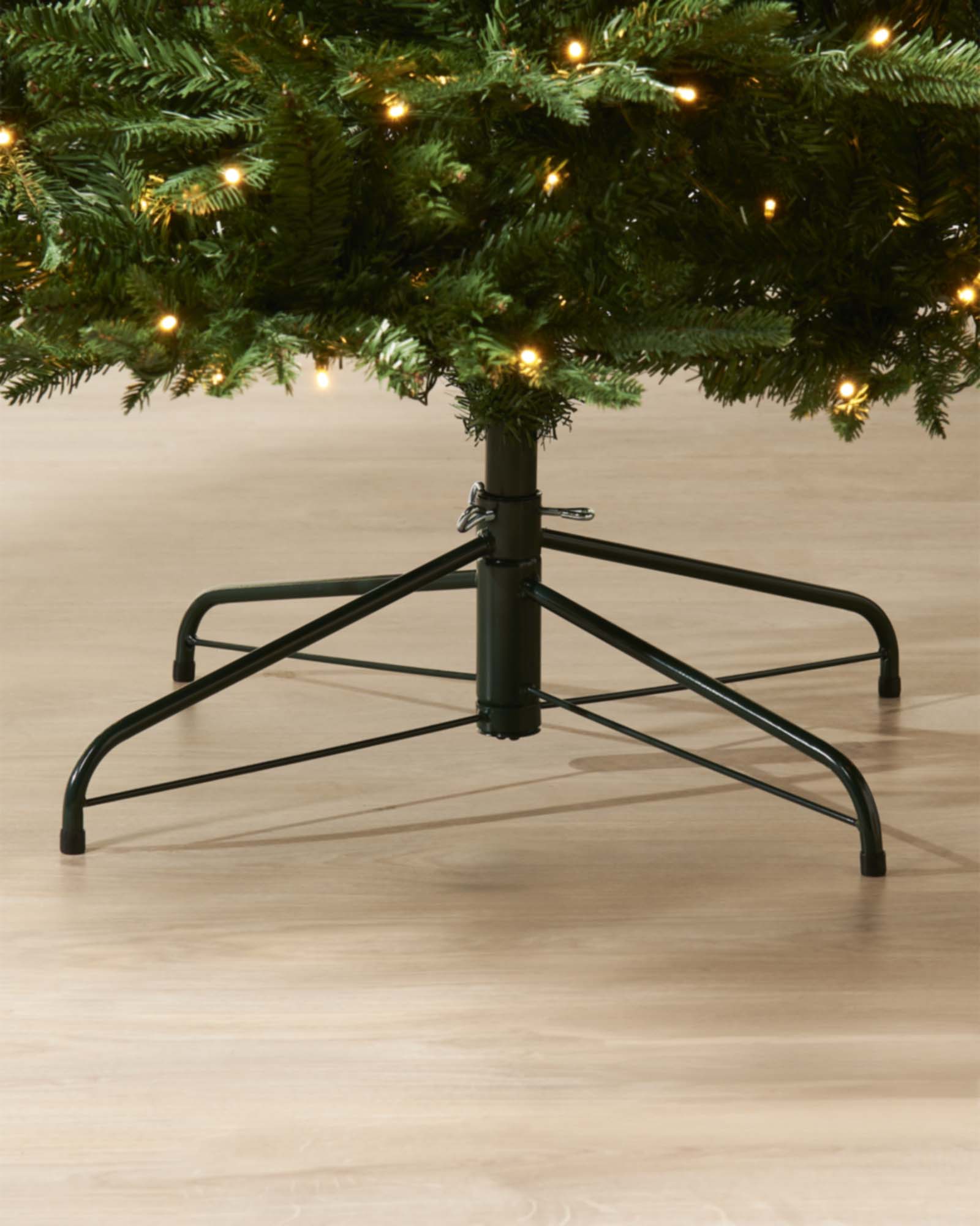 Christmas tree stand with a decorated tree on a wooden floor. Pre-lit Grand Alaskan Fir artificial Christmas tree with warm white LED lights, full mixed PE and PVC branches, displayed against a white paneled wall.