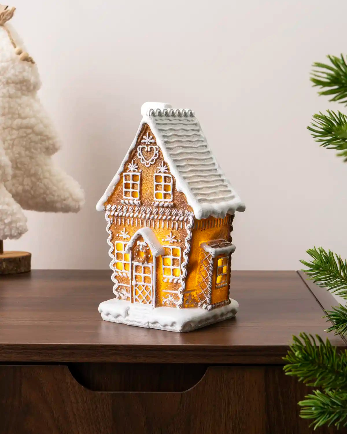 Pre-lit gingerbread house displayed on a wooden cabinet, viewed from an angle with glowing windows, white icing details, and Christmas tree branches framing the scene