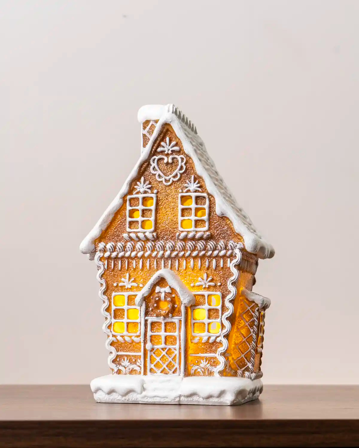 Pre-lit gingerbread house with white icing details, glowing windows, a snow-covered roof, and a snowy base, shown on a wooden tabletop
