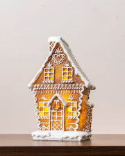 Pre-lit gingerbread house with white icing details, glowing windows, a snow-covered roof, and a snowy base, shown on a wooden tabletop
