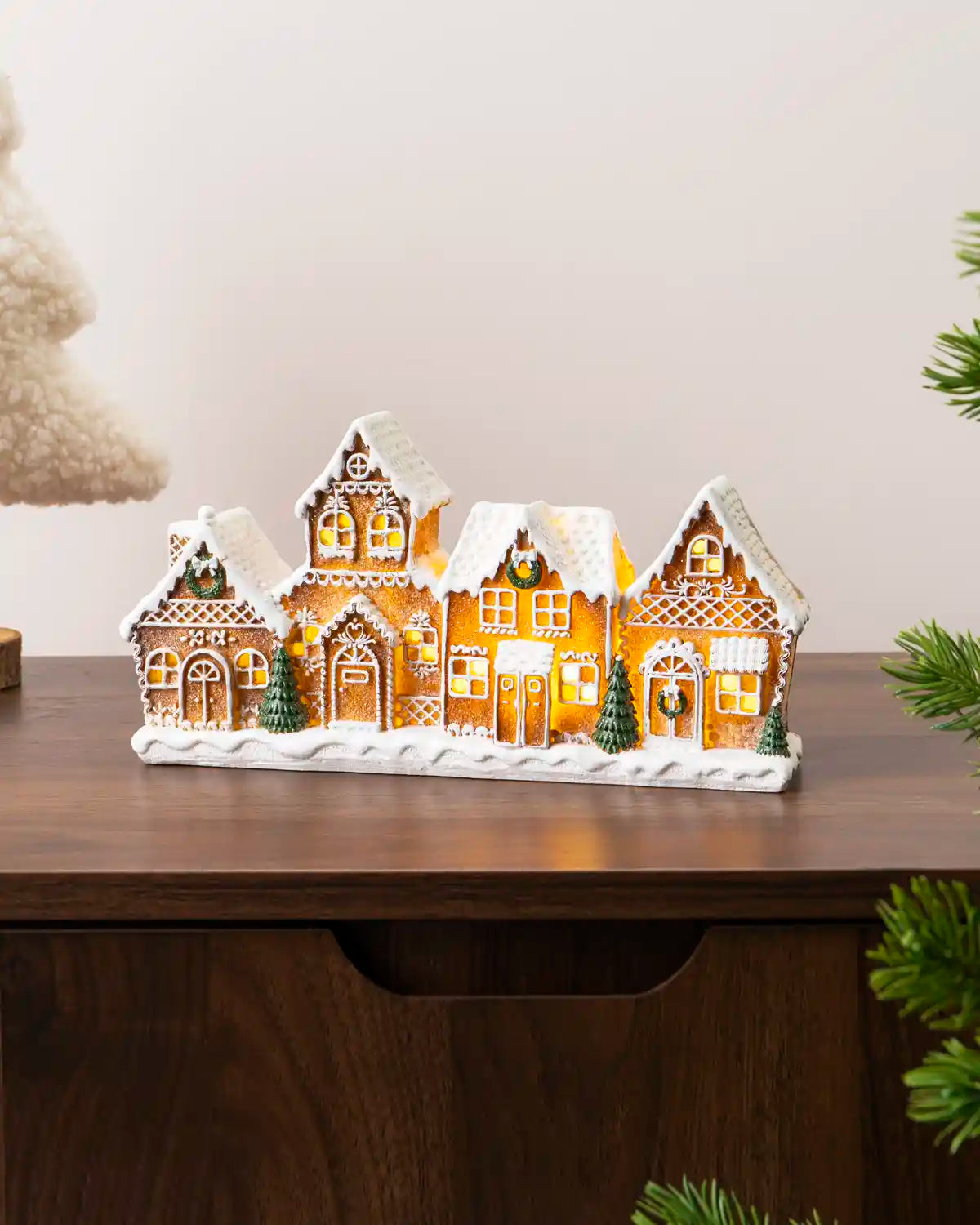 Pre-lit gingerbread village displayed on a dark wooden console, featuring five detailed gingerbread houses with snowy roofs, glowing windows, green wreaths, and small Christmas tree branches around the scene