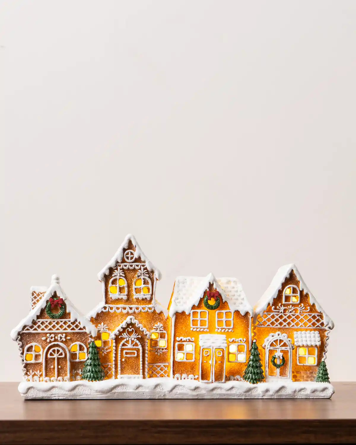 Pre-lit gingerbread village featuring five snow-covered gingerbread houses with glowing windows, white icing details, green wreaths, and miniature Christmas trees on a snowy base
