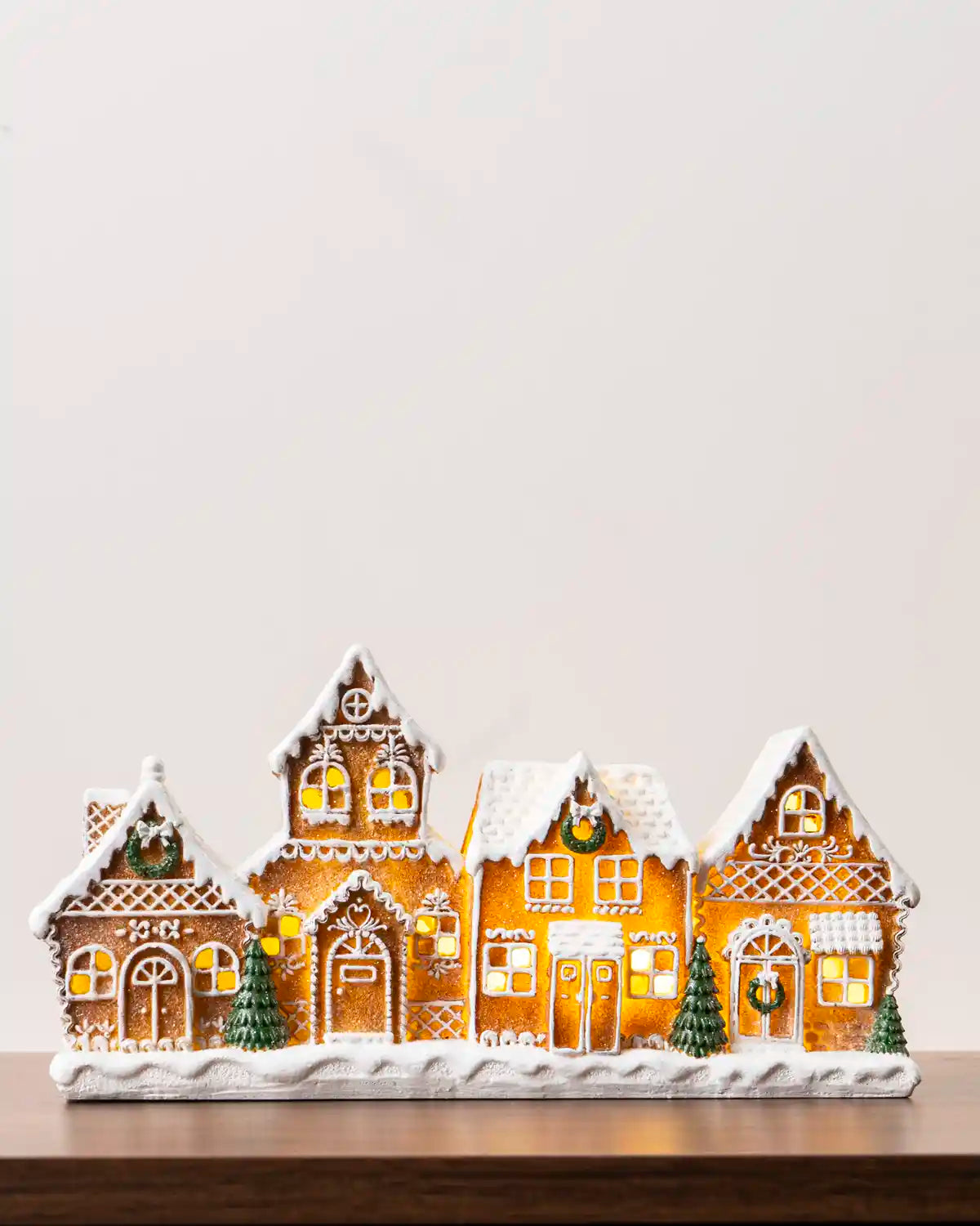Pre-lit gingerbread village featuring five snow-covered gingerbread houses with glowing windows, white icing details, green wreaths, and miniature Christmas trees on a snowy base