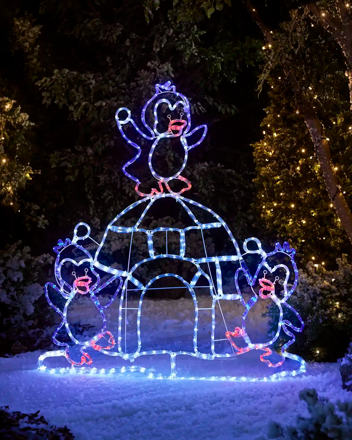 Pre-lit penguin igloo rope light silhouette glowing in blue and white lights outdoors on snow, featuring three penguins surrounding an igloo