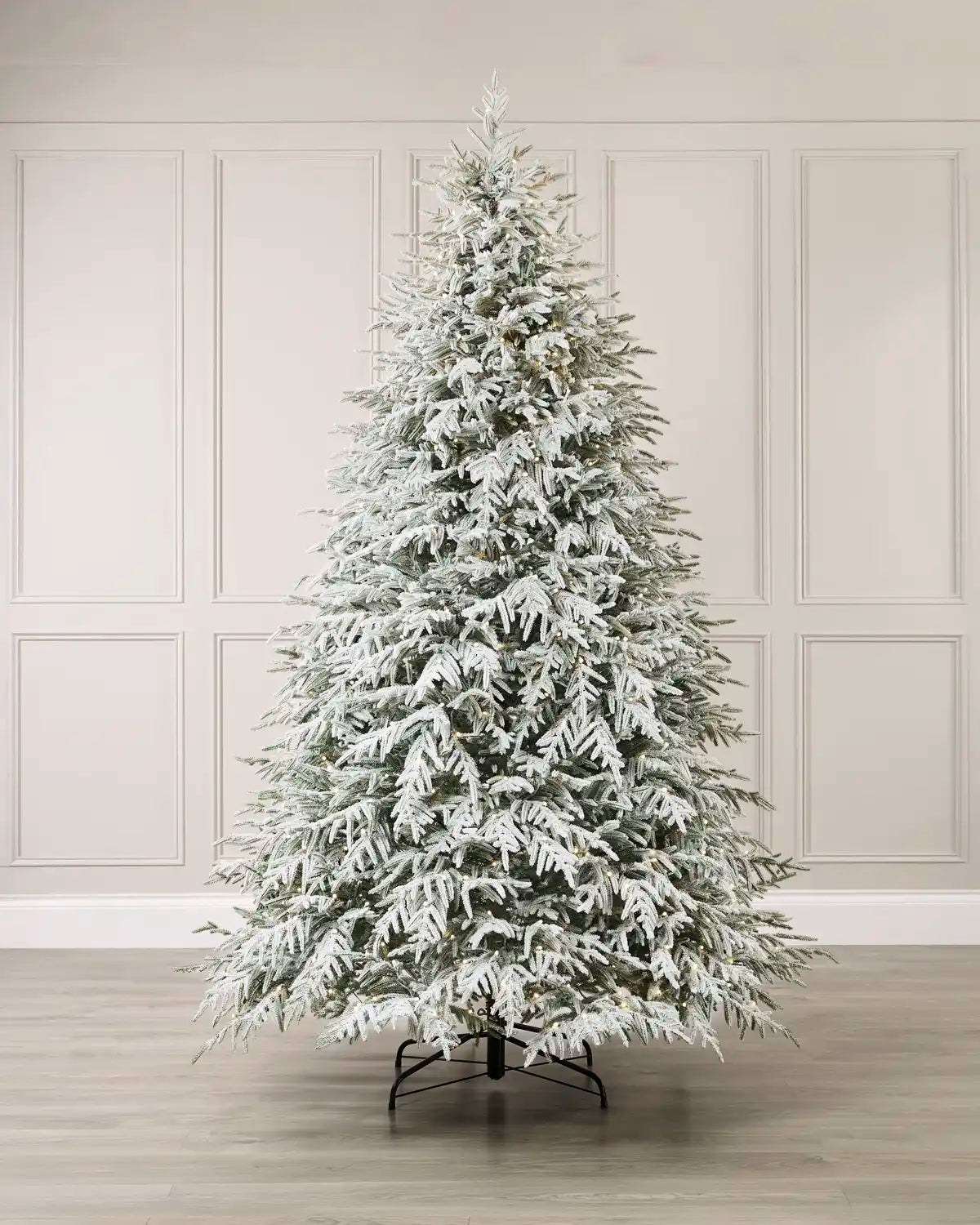 Pre-lit snow-flocked Grand Duchess Christmas tree, 7.5 feet tall, covered in snowy white flocking and warm white lights, displayed against a white paneled wall