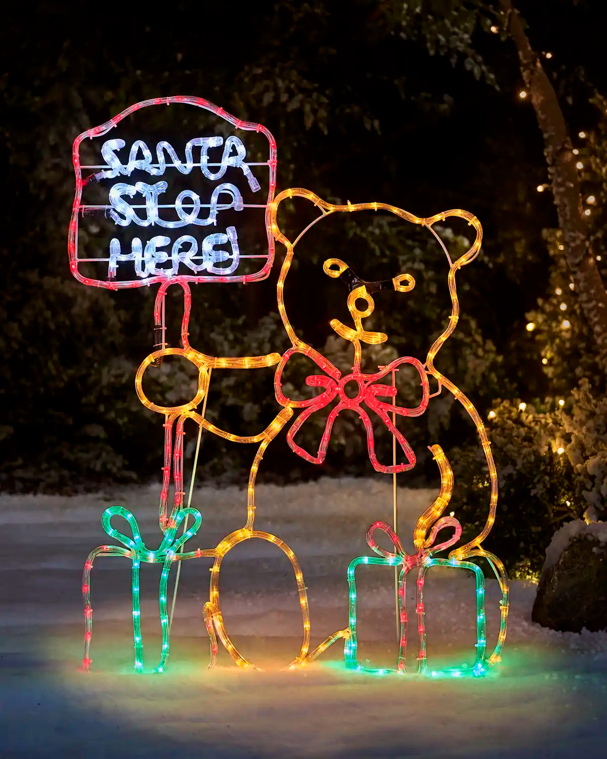 Pre-lit teddy bear Christmas silhouette holding a “Santa Stop Here” sign, with a large red bow and two illuminated presents at its feet, displayed outdoors in a snowy setting